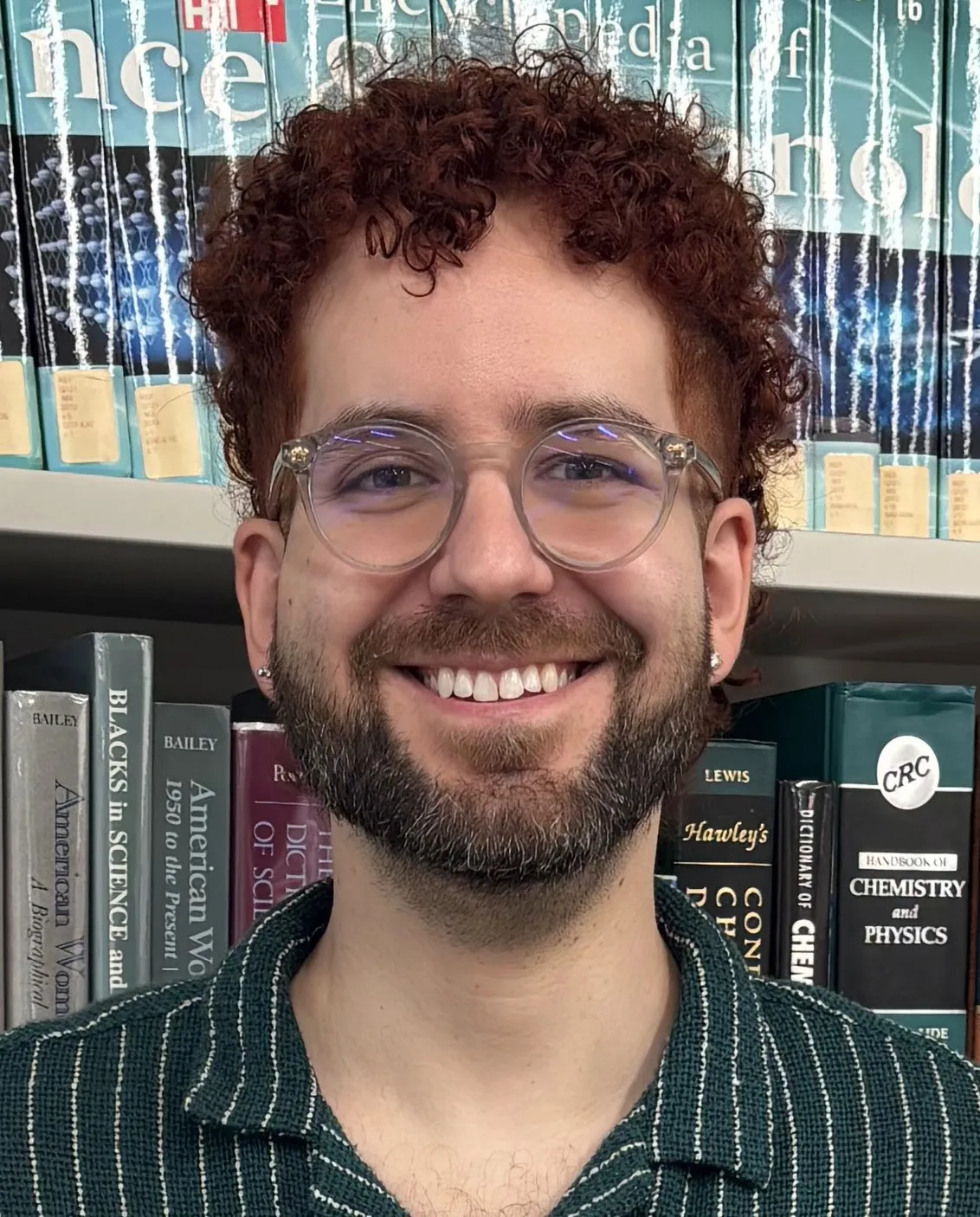 Jorge Enrique Cartaya, smiling, wearing round clear-framed glasses and a dark green striped shirt, in front of library shelves.